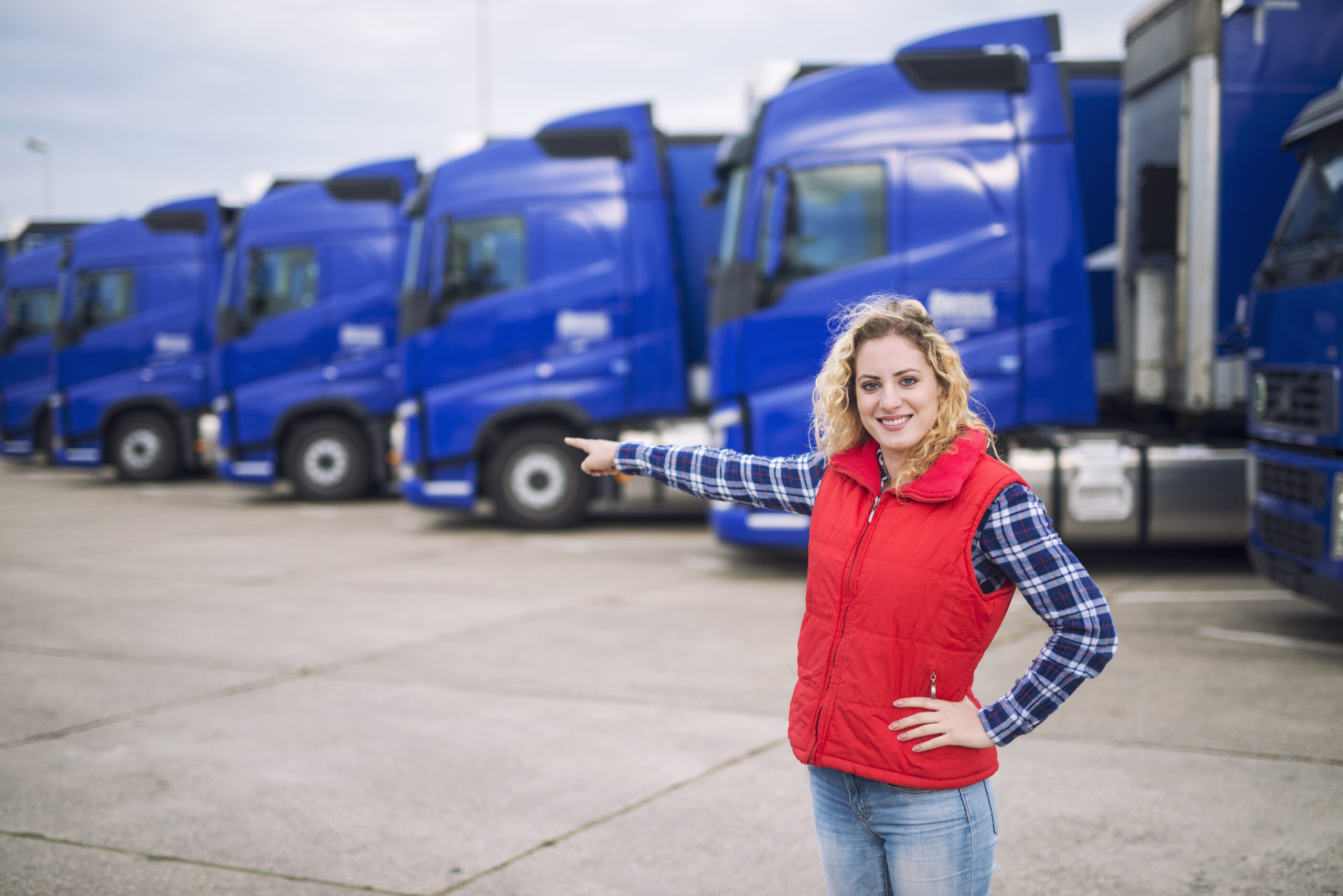 Female trucker standing in front of parked trucks and pointing her finger to the transportation vehicles.